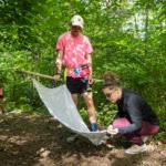 Professor Erin Heller knees, looking for ticks on the cloth Ethan Caldwell is dragging across the ground to collect ticks. They are standing on a trail, surrounded by trees.