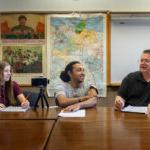 Professor Gerry Sherayko, Tristan Gregory '25, and Maegan Lloyd '26 sit at a table, with papers in front of them and an iPhone on a tripod, which they use for their project interviews.